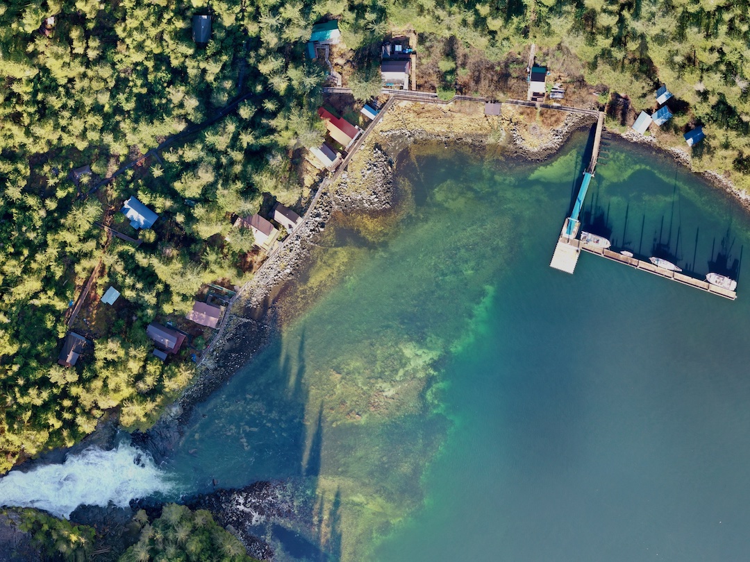 Aerial orthographic view
of Baranof Warm Springs. There's a waterfall in the bottom left of the frame
and a dock with boats with shadows streaming upwards as the sun shines from
the south. A boardwalk runs across the waterfront and there are quite a few
houses.